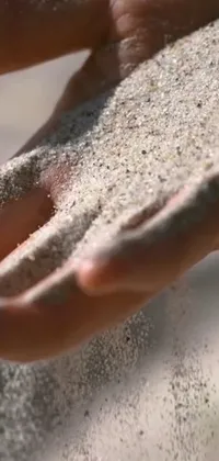 This stunning live wallpaper for your phone features an up-close shot of sand grains in someone's hands, with clear and detailed imagery, captured in beautiful daylight, from an attractive angle