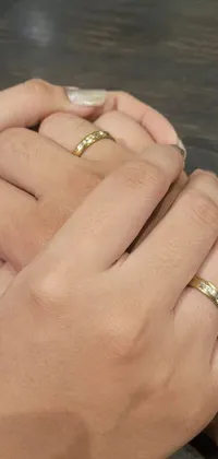 This phone wallpaper features a close-up of two people holding hands, showcasing two gold rings