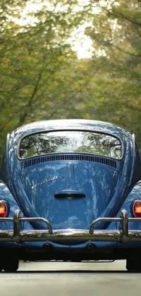 This lively phone wallpaper features a vintage blue car with a rustic tail fin design driving down a scenic road with tall trees on either side