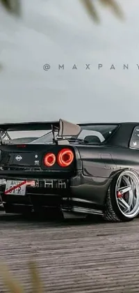 This live wallpaper for your phone showcases a black sportscar, the Nissan GTR R 3 4, parked next to a serene body of water