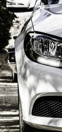 This live wallpaper features an up close image of a glossy white metal car with its headlights on, parked by the side of the road
