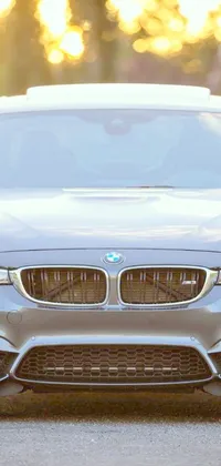 This stunning phone live wallpaper showcases a shining, silver BMW parked on the side of a beautifully scenic road, with trees and rolling hills in the background