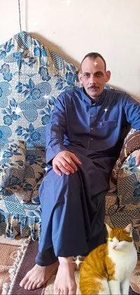 This phone live wallpaper showcases a sophisticated man in traditional dau-al-set clothing, sitting on a cozy couch in a blue-hued living room