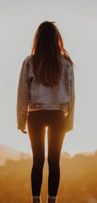 This live phone wallpaper features a woman standing on a large rock, backlit by the sun, wearing a jean jacket