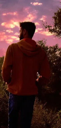 This phone live wallpaper features a man wearing a pink hoodie standing on a cement wall in a forest at sunset