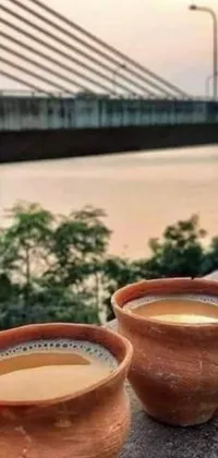 This live phone wallpaper showcases an invigorating scene of two cups of coffee adorning a wooden balcony facing a stunning bridge backdrop in an idyllic setting evoking an Assam tea garden