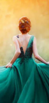 This live wallpaper features a stunning woman in a green dress sitting in a field with her back turned, set against a beautiful sunset