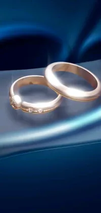 This live wallpaper depicts two digital wedding rings shining with a metallic sheen on a blue background