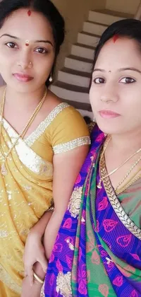 This colorful phone live wallpaper features two beautiful women dressed in vibrant Indian saris, posing for a picture or Instagram post