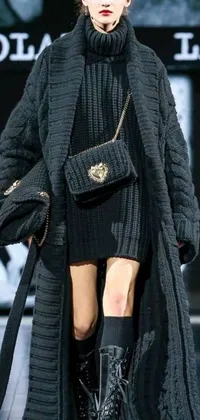 This phone live wallpaper features a fashionable woman strutting down a runway in a black coat, paired with an oversized sweater and Versace handbag