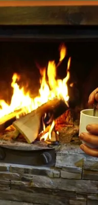 This phone live wallpaper features a realistic photograph of a person holding a cup of hot beverage in front of a cozy fire