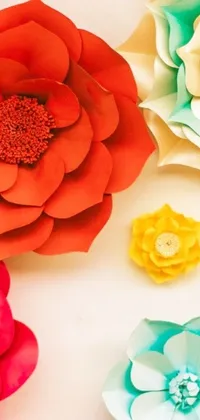 Get lost in the beauty of a phone live wallpaper featuring bright and colorful paper flowers arranged on a table with a blurred garden background in shades of red, teal, and yellow