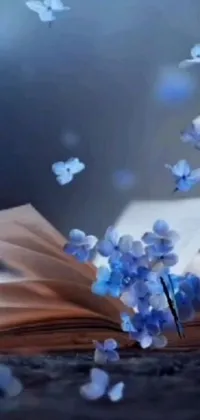 This stunning phone live wallpaper features an open book on a wooden table surrounded by flying books and blue petals