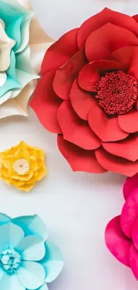 This phone live wallpaper showcases a bunch of paper flowers in red, teal and yellow arranged on a table with large, opaque blossoms