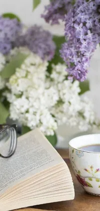 This stunning phone live wallpaper depicts an open book and a cup of coffee on a wooden table, accompanied by lilacs and black-rimmed glasses