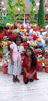 This phone live wallpaper showcases two girls posing in front of a colorful flower stand that amplifies the celebratory atmosphere