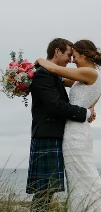 This stunning live wallpaper captures a romantic moment between a man in a traditional kilt and a woman in a gorgeous wedding dress standing by the ocean