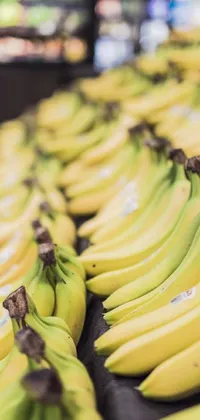 This phone live wallpaper features a vibrant bunch of bananas resting on a wooden table in a bustling supermarket