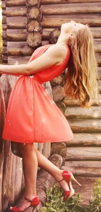This phone live wallpaper showcases a stunning woman wearing a vibrantly coral red dress, accentuated by red high heels, leaning gracefully against a rustic fence