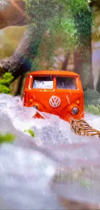 This phone live wallpaper showcases a bright orange Kombi van driving through a lush forest filled with towering trees