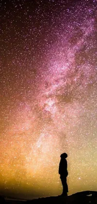 Invite serenity and awe to grace your phone screen with this beautiful live wallpaper featuring a small silhouette standing atop a hill against a backdrop of milkyway light and a sky full of stars