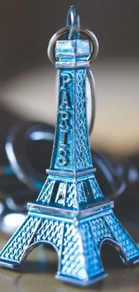 Looking to personalize your phone wallpaper? Check out this stunning live wallpaper featuring a keychain shaped like the Eiffel Tower! Captured beautifully in close-up by the talented Raphaël Collin, this metallic blue keychain is a feast for your eyes and is adorned with chrome hearts for contrast