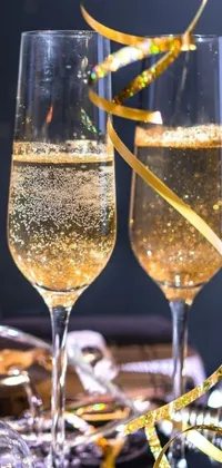 This phone live wallpaper features two glasses of champagne on a table, decorated with golden ribbons and metallic flecks, as bubbles rise from the glasses