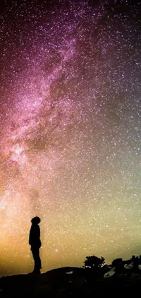 This intense phone live wallpaper showcases a lone person standing atop a hill, gazing up at a mesmerizing, multicolored sky full of stars