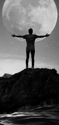 This stunning phone live wallpaper showcases a man standing atop a rocky formation under a full moon, in a black and white photograph