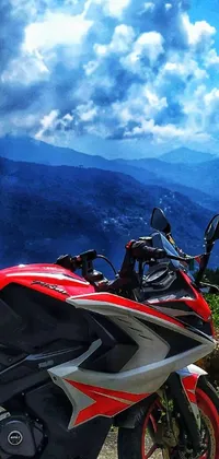 This live wallpaper depicts a red and black motorcycle parked on the side of a beautiful winding road that cuts through a majestic mountain range