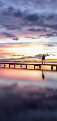 This phone live wallpaper features a man standing on a pier by the water with a romantic and colorful sunset in the background