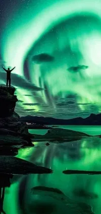 This live phone wallpaper depicts a serene scene of a person standing on top of a rock near a sparkling body of water, in stunning aurora green