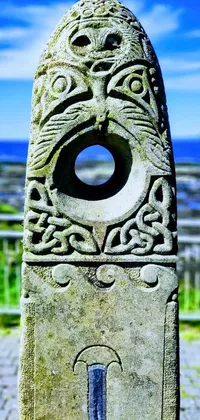 This phone wallpaper displays a close-up of a intricately carved stone object with Celtic designs near a seashore