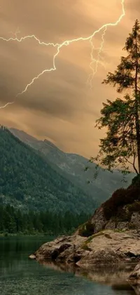 This live wallpaper for your phone features a powerful yet serene scene: a tree standing on top of a rock by a body of water, with lightning behind it and a mountain forest in the background