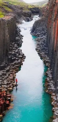 This phone wallpaper is a stunning optical illusion featuring a man standing on a cliff overlooking a vibrant river