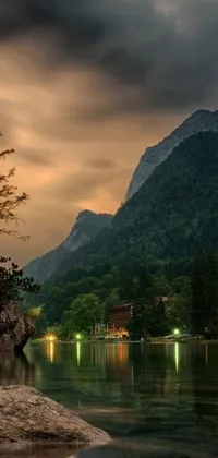 This Live Wallpaper features a serene body of water reflecting the soft colors of an evening sky, with a majestic mountain in the background, hidden village, and lush trees and cliffs