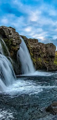 This live wallpaper features a tranquil waterfall with a large rock formation in the background and a grassy area in the foreground against a blue sky