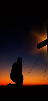 This phone live wallpaper features a beautiful and serene image of a man kneeling at the foot of a wooden cross during sunset