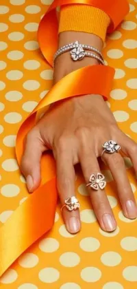 This engaging phone wallpaper showcases a close-up image of a hand delicately holding an orange ribbon against a background of diamonds and clovers