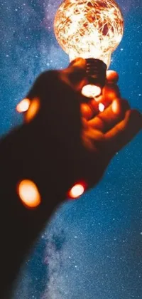 This live wallpaper for your phone depicts a fascinating and awe-inspiring image of a hand holding a light bulb amidst a background of space and light