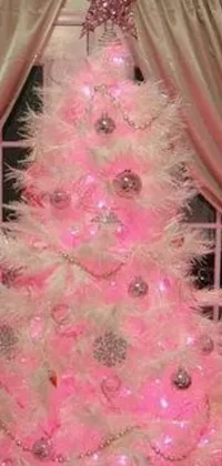 This phone live wallpaper features a vibrant pink Christmas tree in front of a window, adorned with sparkling lights, a feather boa, and a crown of white lasers