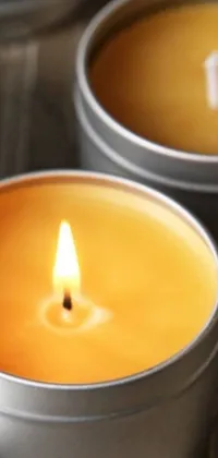 This phone live wallpaper features a highly detailed close up of two tins of candles on a table, with a warm yellow light emitting from the flickering flames
