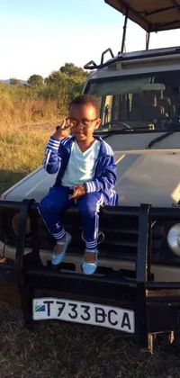 This phone live wallpaper features a young boy wearing a tracksuit, sitting on the bumper of a vehicle on the savanna