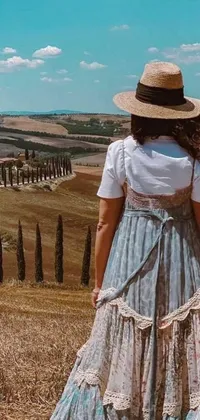 This phone live wallpaper features a picturesque scene of a woman in a flowing dress and a hat standing in a field surrounded by rolling hills in Tuscany