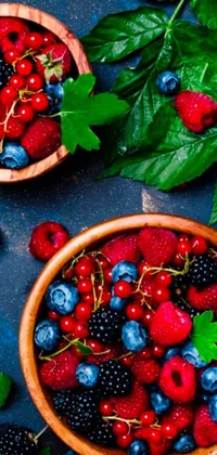 This phone live wallpaper showcases a wooden bowl filled with fresh blueberries and raspberries, surrounded by plates of fruit, set against shifting backgrounds of blue skies and lush green forests