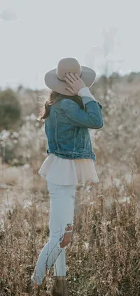 This unique live wallpaper showcases a serene image of a woman wearing a denim jacket and white hat standing amidst a vast field of tall grass