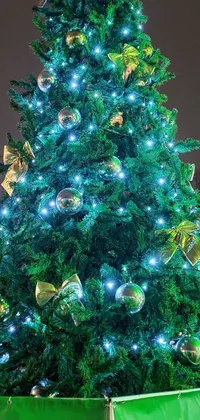 This live wallpaper features a large Christmas tree adorned with glittering lights, shining baubles, and golden ribbons set against a dark night sky