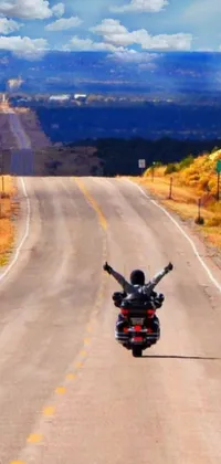 This dynamic phone wallpaper features an exhilarating scene of a man riding on the back of a motorcycle down a road in New Mexico