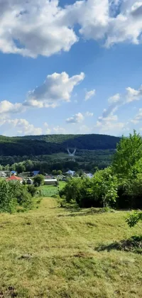 This phone live wallpaper depicts a peaceful scene with a cow grazing on a green field, a small town in the foreground, and hills in the background