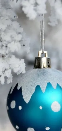 This phone live wallpaper features a beautifully designed blue and white Christmas ornament hanging from a well-decorated tree, set against a serene winter landscape
